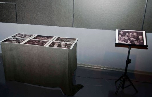 Mesa cubierta con una tela gris que sostiene varias fotografías en blanco y negro dispuestas en fila; a la derecha, un atril con una imagen similar también en blanco y negro, mostrando a varias personas. La escena está ambientada en una sala de exposición con iluminación tenue.